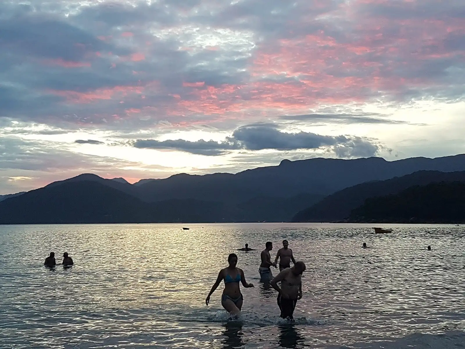 Fim de tarde na Praia do Lázaro: gente entrando no mar calmo, a serra escura e o céu rosado